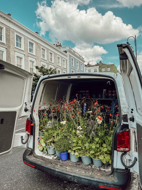 The open rear of a white delivery van filled with potted plants and flower arrangements, including poppies, daisies, and other flowering plants in various white and blue containers. The plants display