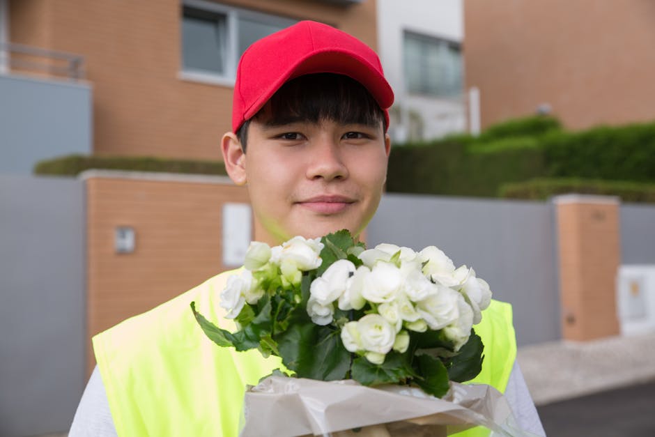 A young delivery person wearing a red cap and high-visibility yellow vest stands outdoors in a residential area, holding a bouquet of fresh, white flowers wrapped in beige paper. The arrangement featu