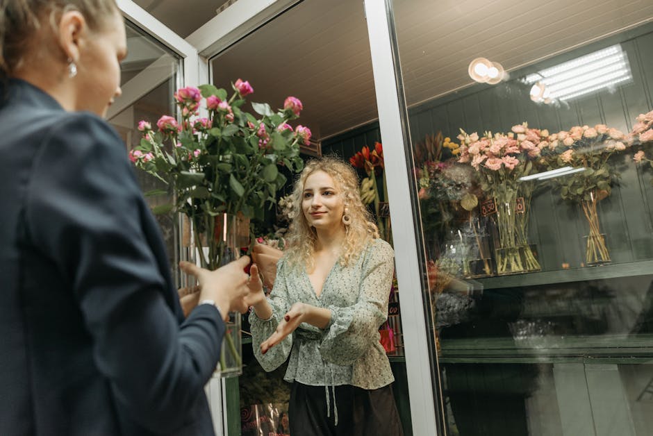 A young woman with curly blonde hair and light makeup receives a large bouquet of bright pink roses with green foliage from a florist at the flower shop. The bouquet is wrapped in simple brown craft p