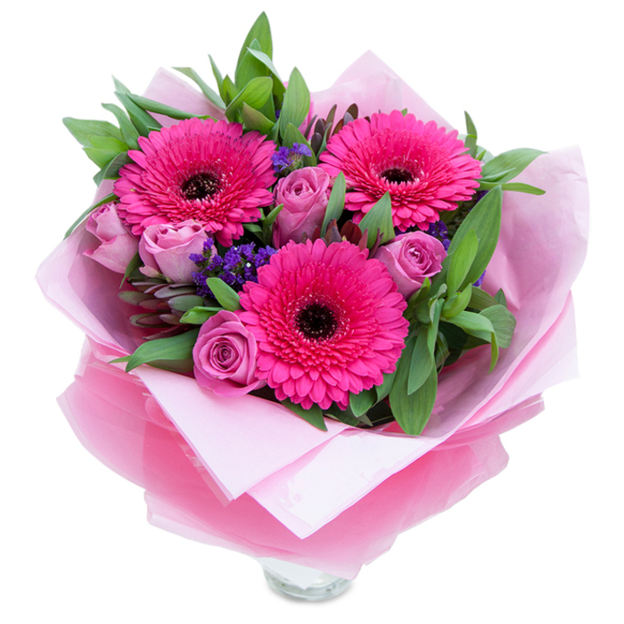 The composition begins with three vivid Gerbera daisies in a clear fuchsia, each blossom open and bright with a contrasting deep centre that grounds the arrangement. Around them, roses in a gentle lavender-pink or dusty rose remain in a budding stage, their layered petals suggesting tenderness and time yet to unfold. tucked between these larger forms are delicate sprays of purple statice that bring a textural lift and a cool, almost wildflower note to the bouquet. Surrounding foliage - broad, slightly waxy leaves reminiscent of salal and the rounder, muted green of eucalyptus - provides a leafy theatre that cradles each bloom and adds a fresh, dewy sense of the garden. The bouquet is presented in an inner sleeve of soft pale-pink tissue and an overwrap of slightly crinkled, translucent fuchsia-pink cellophane that shimmers when caught by morning or late-afternoon light; the wrapping is folded deliberately so the arrangement reads as both casual and refined. A clear glass vase rim peeks out at the base, indicating the flowers are arranged with longevity and immediate display in mind. The scent is subtle - a light rose perfume layered with the clean, green notes of fresh foliage - and the overall mood is one of gentle celebration: perfect for a birthday in a Bloomsbury flat, a small anniversary dinner near Drury Lane, or a spontaneous gift picked up from a stall at the Covent Garden Market. It's a bouquet designed to lift the room with colour and calm.
